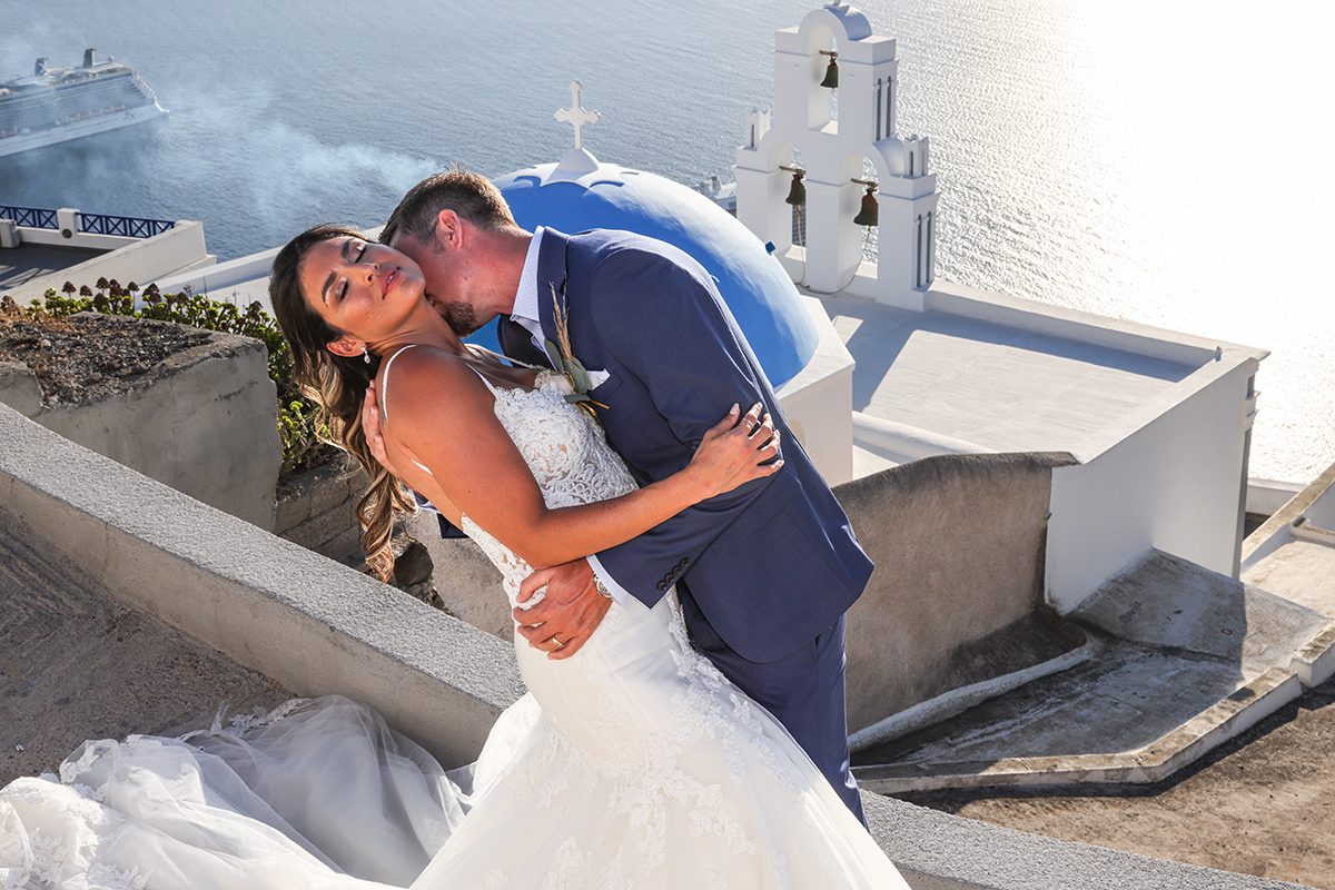Newlyweds kissing above the Three Bells of Fira blue dome with the Aegean shimmering behind