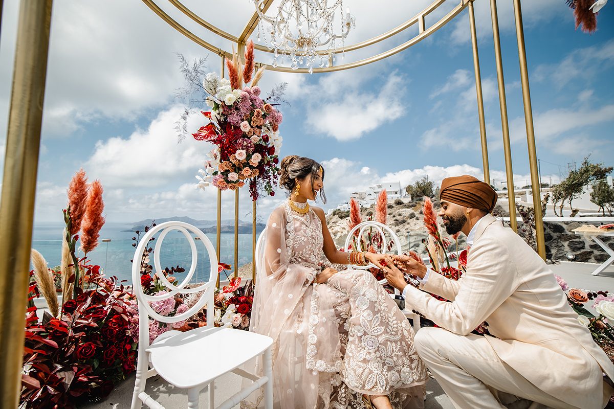 Couple exchanging rings under a gold gazebo dressed with red and pink flowers above the caldera