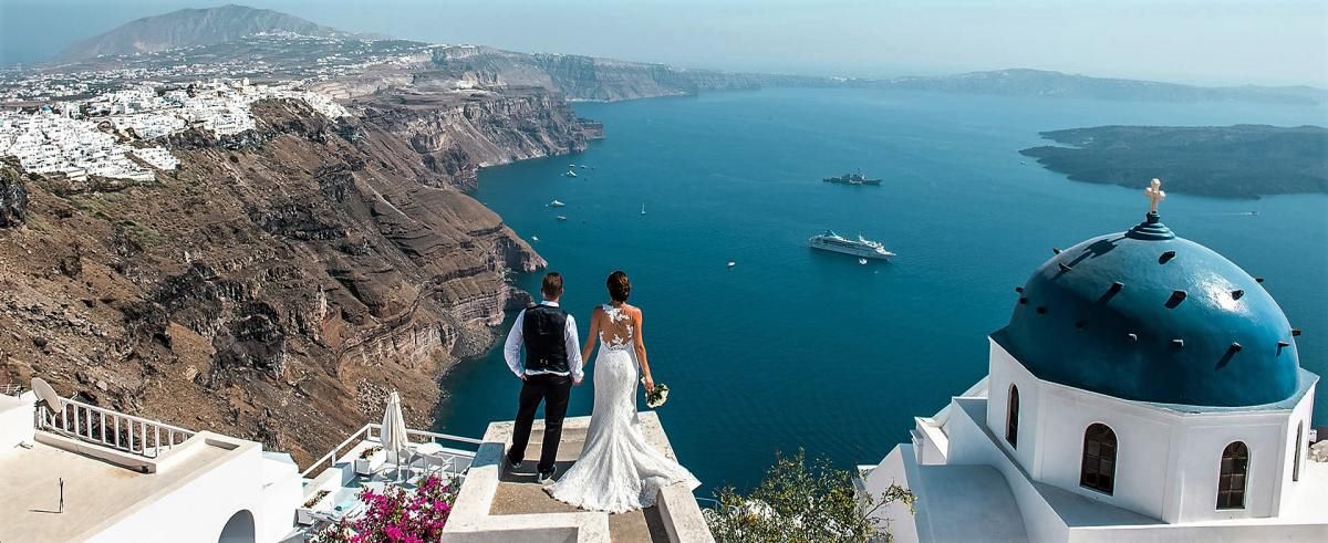 Bride and groom gazing over the Santorini caldera beside a blue-domed church