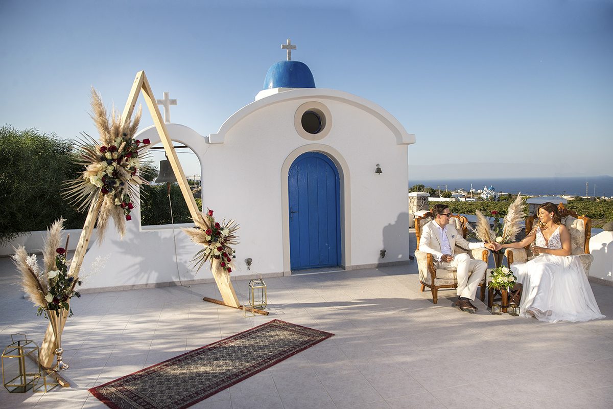 Wedding ceremony set at a blue-domed Santorini chapel with a boho arch and the sea beyond